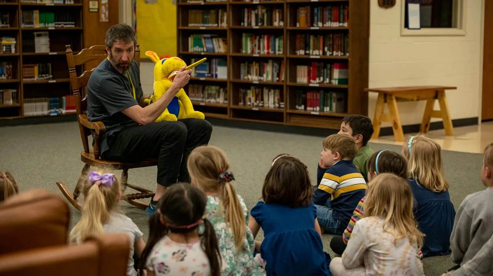 A teacher reading aloud from a rocking chair to lower school students seated on the library floor