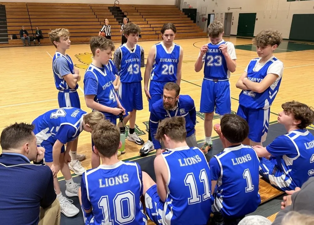The Eukarya Lions boys basketball team gathered around their coach during a timeout, in blue jerseys on the home court
