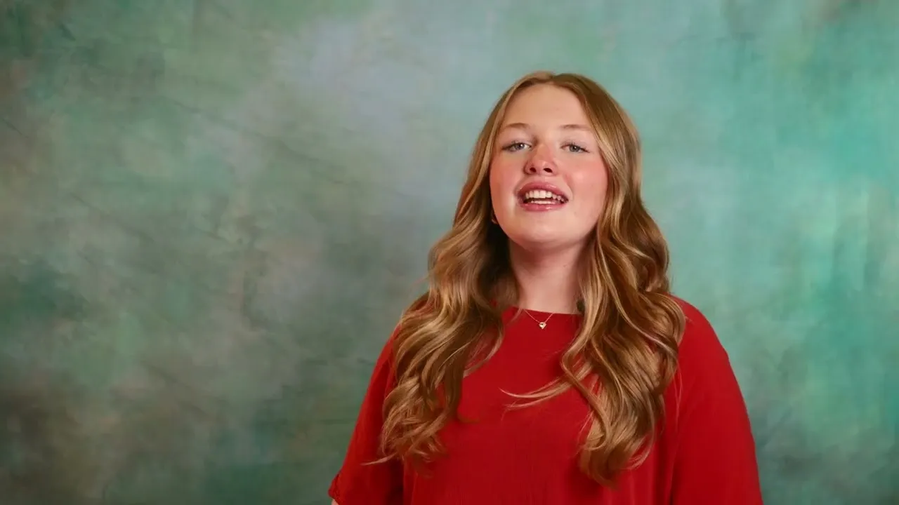 An upper school student in a red top speaking to camera against a mottled green studio backdrop