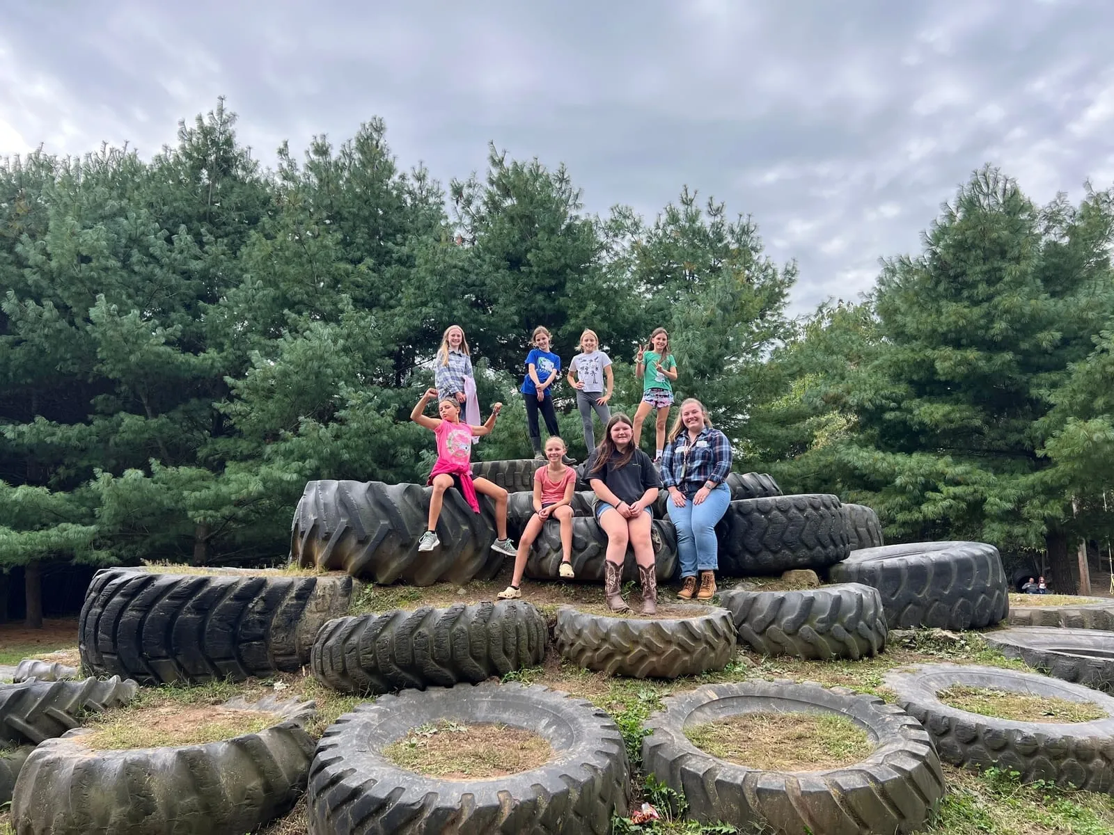 Students perched together on the tractor tyres at the edge of the field, pines behind them