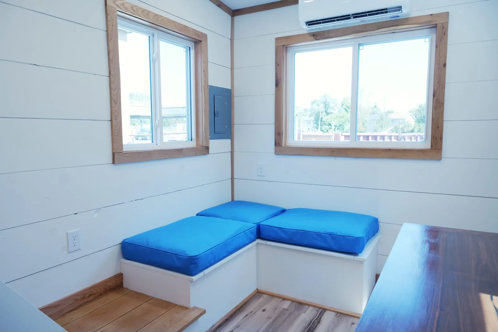 L-shaped white storage bench with royal-blue cushions beneath two wood-trimmed windows, with the mini-split head mounted above
