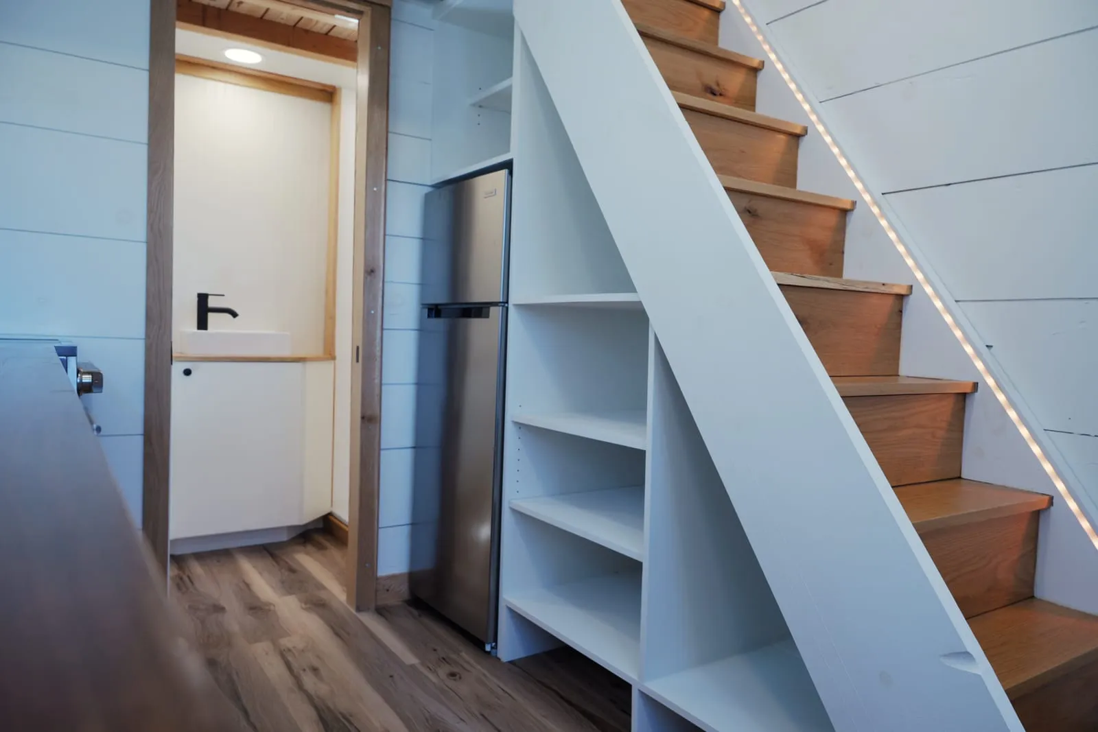 Oak staircase with warm LED strip lighting, built-in white shelving underneath and a stainless refrigerator alongside