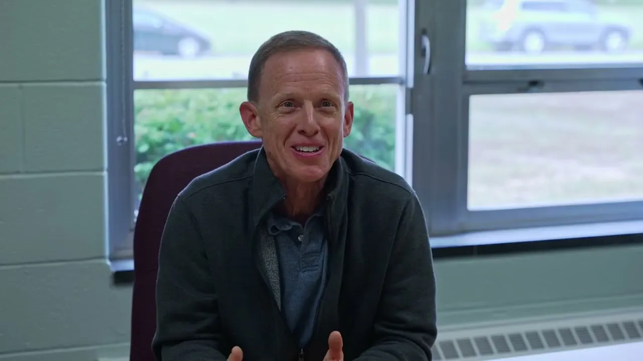 A teacher seated at a desk by a bank of classroom windows, speaking to camera